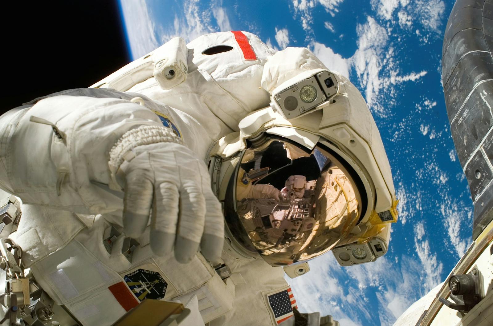 This picture shows an american astronaut in his space and extravehicular activity suite working outside of a spacecraft on a human spaceflight mission. In the background parts of a space shuttle are visible. In the far background of the picture planet earth with it's blue color and white clouds is shown as well as a patch of black space.